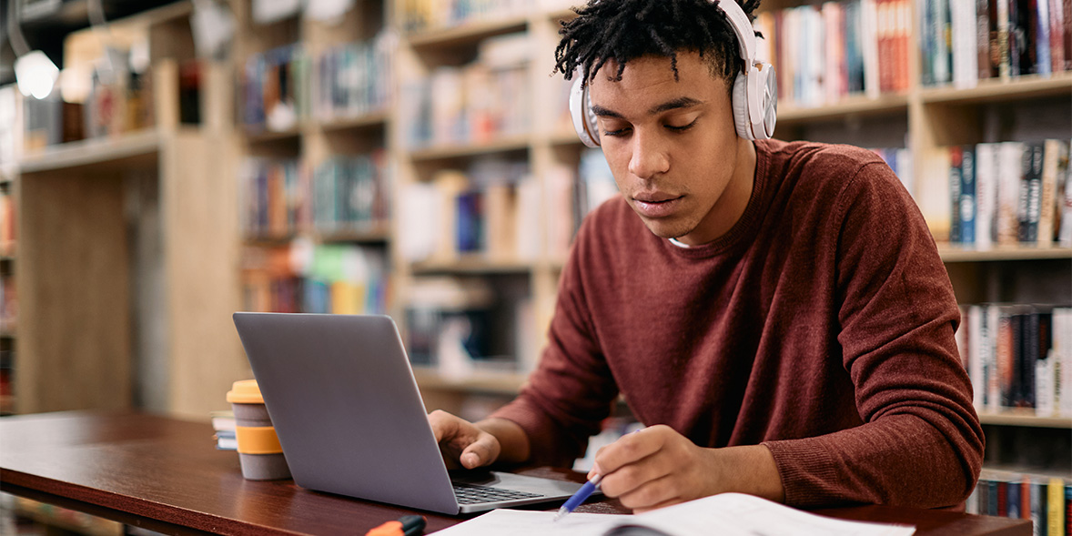 Man using a laptop in a library with a notepad, pen and a travel cup.